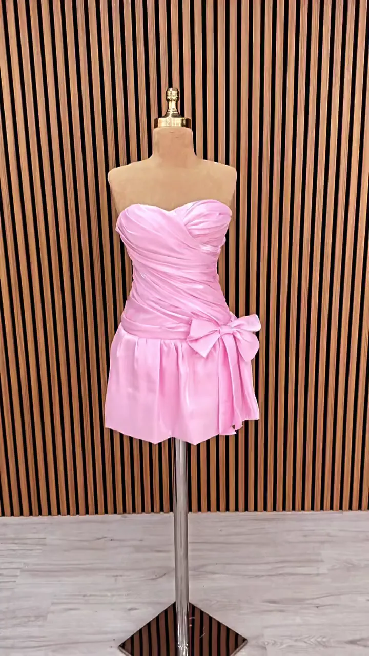 Kalp yaka düşük bel yan fiyonklu saten mini after party elbisesi, açık pembe renkte ahşap çıtalar önünde önden.